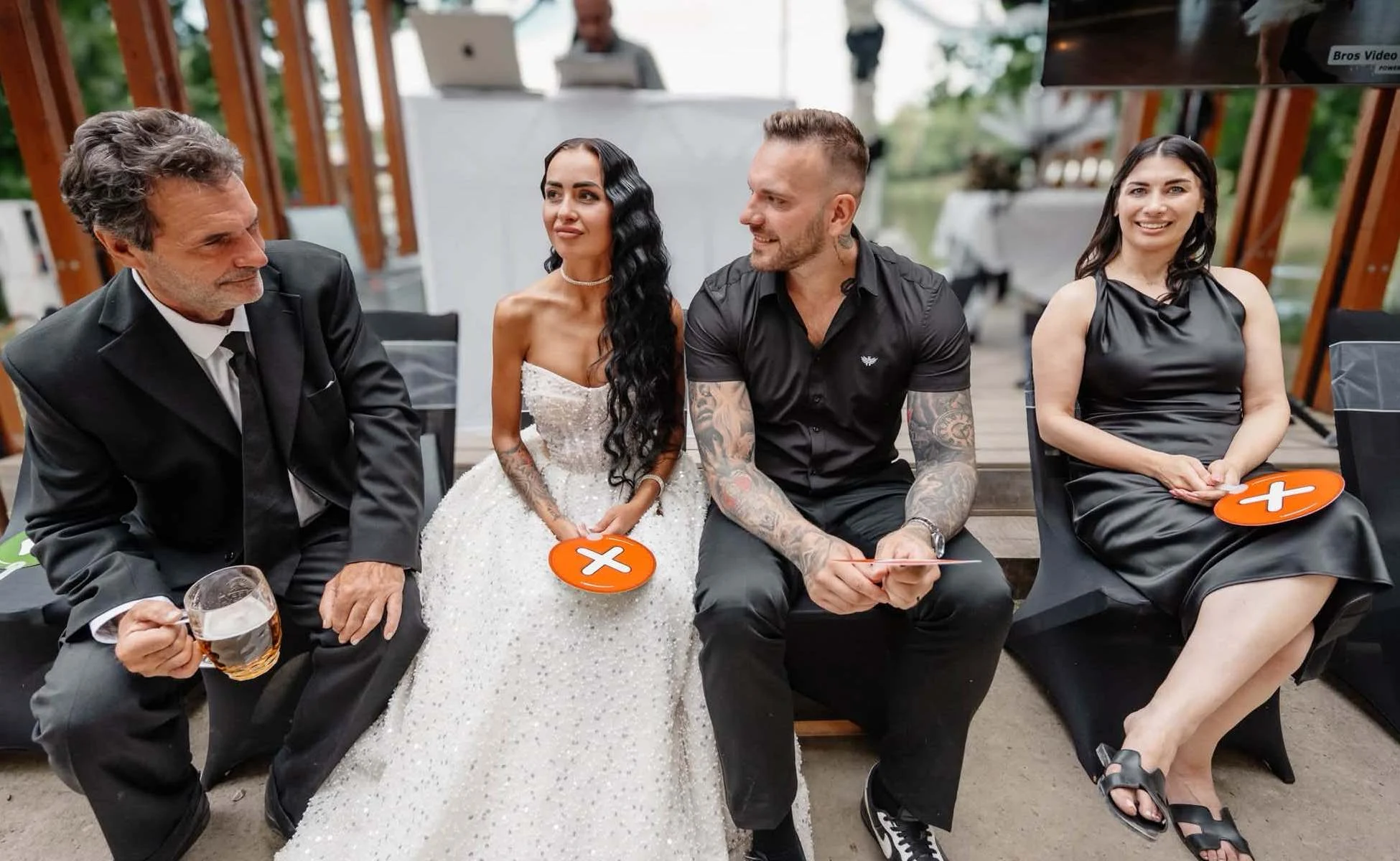 Hvězdná porota Cover StarDance na svatbě Usměvavá svatební porota složená z novomanželů a rodiny s hodnotícími cedulkami v rukou během zábavné taneční show Cover StarDance. V pozadí je připravený DJský pult a projekční obrazovka.