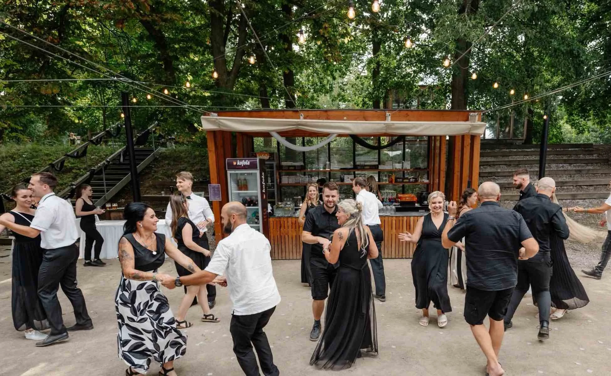 Taneční soutěž Cover StarDance na svatbě Skupina usměvavých svatebních hostů tančí v párech na venkovním parketu pod světelnými řetězy během autorské zábavné show Cover StarDance.